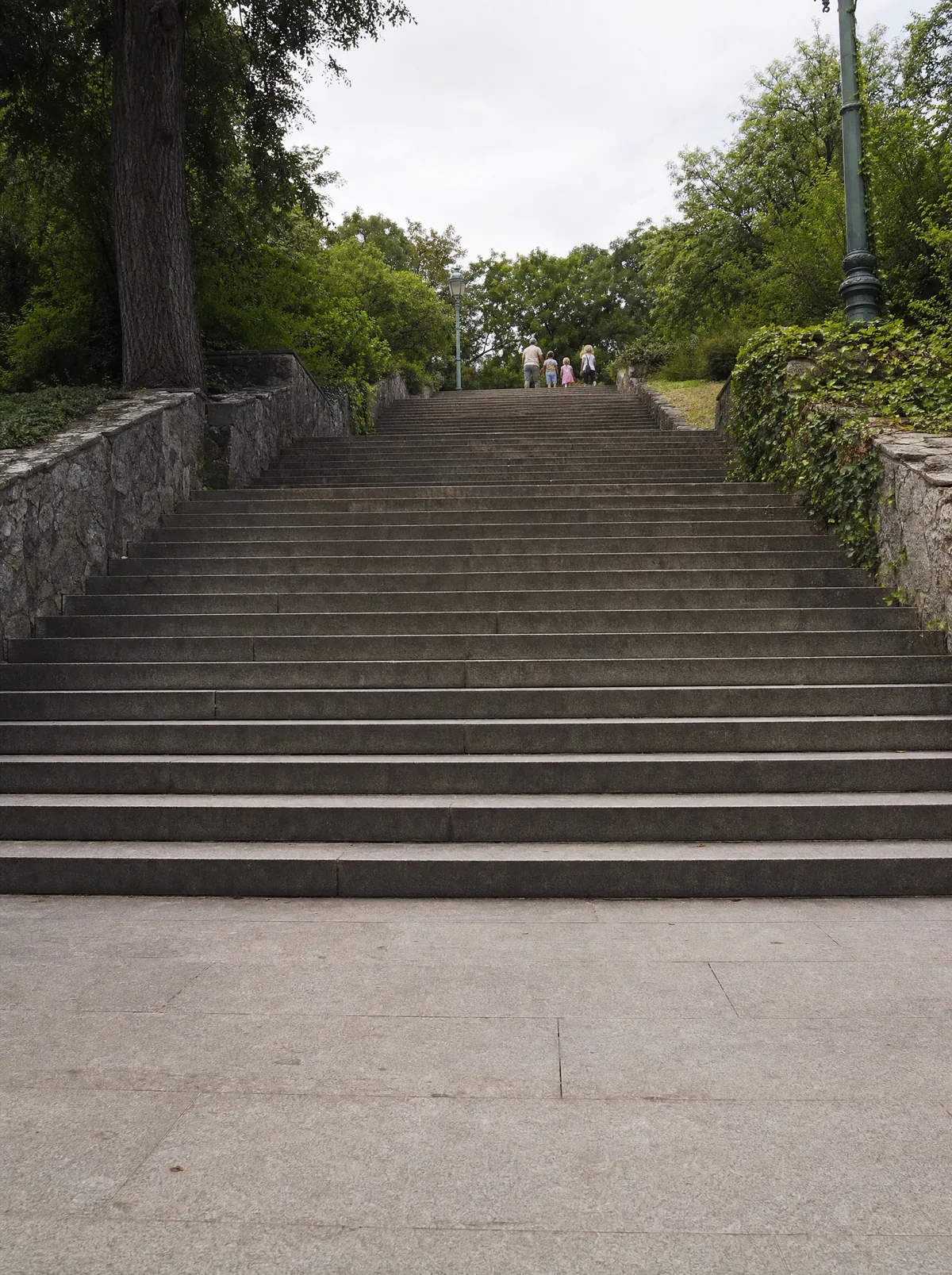A flight of steps seen from the bottom, nobody on them, a family already at the top. Read at a distance so the frame is about being apart from something. Never a fall, never an injury, never a hospital, never anyone being caught.